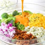 Pouring buffalo sauce over Buffalo Broccoli Salad ingredients in a clear glass bowl on a white table