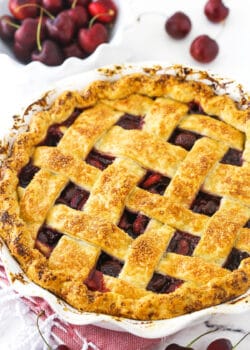 A cherry pie inside of a 9-inch pie dish with a bowl of sweet cherries in the background