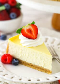 A slice of homemade cheesecake with whipped cream and berries with a bowl of berries in the background