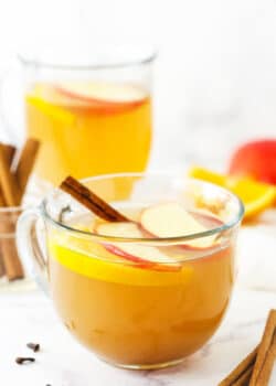 A mug of hot apple cider with a jar of cinnamon sticks and another mug of cider behind it