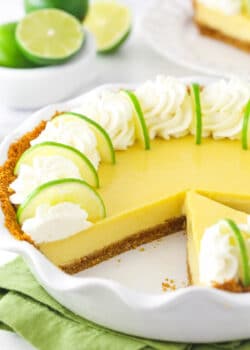 A homemade key lime pie inside of a pie dish with one slice missing and a bowl of fresh limes in the background