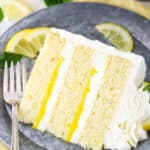 A slice of Lemon Mascarpone Layer Cake on a metal colored plate with a fork and cut lemons in the background