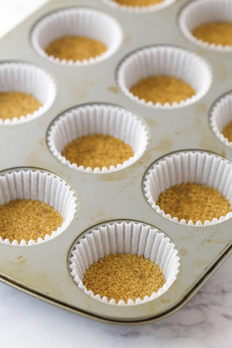 Graham cracker crust pressed into the bottoms of cupcake liners ready to be baked.