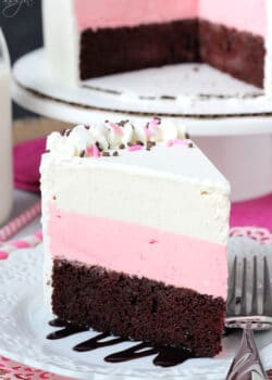 A slice of chocolate, strawberry and vanilla cake on a plate with the remaining cake on a cake stand behind it