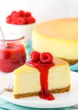 Slice of New York Style Cheesecake topped with raspberry sauce and raspberries on a white plate with a fork.