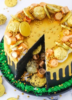 Overhead view of a Pot Of Gold Cake with a slice taken out and decorated with gold wrapped chocolate coins and gold drip in a black pan