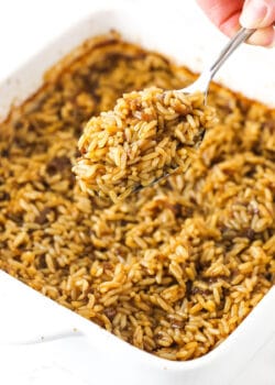 A metal spoon holding up a big bite of baked rice with a pan full of more rice beneath it